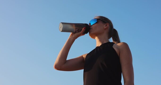 thirsty woman drinking water from bottle against blue sky background after outdoor workout. body hydration