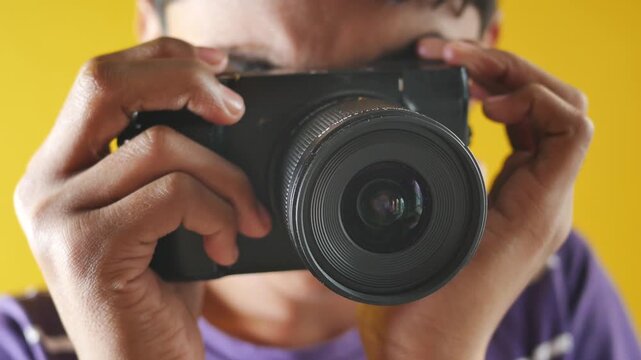Close up shot of person holding camera and taking picture with lens focused on viewer against yellow background