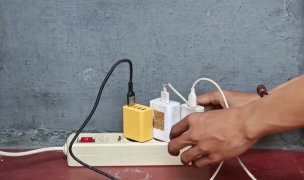 Multiple USB wall chargers connected to a power strip with charging cables against a gray concrete wall. Concept of electricity, device charging, home power, and technology.