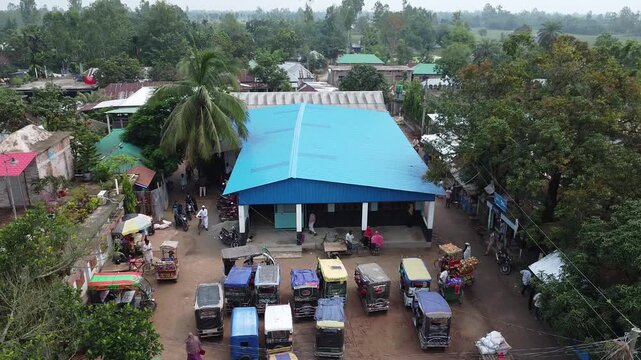 Aerial view of a rural marketplace featuring a large blue-roofed pavilion and various motorized rickshaws parked in the sandy courtyard during the day market village rickshaw transport street