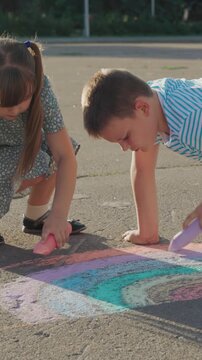 Boy and a girl are playing with chalk on the pavement. The boy is holding a pink and blue stick