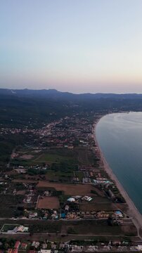 An expansive panoramic aerial drone shot showcasing the coastal settlement of Acharavi and Almyros, nestled beautifully between the long sandy shore and the lush green hills of northern Corfu Island,