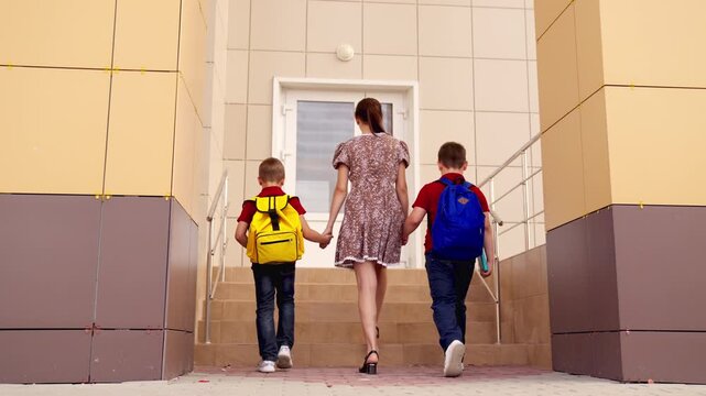 Mom accompanying boys to school, Holding hands for safety, Backpacks ready for class, School entrance, Parental care, Children prepared for lessons, Modern school facade, Boys with colored bags