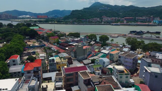 Aerial flyover reveals Coloane Village historic rooftops beside river channel with Zhuhai mountain cable car and Cotai resorts on horizon