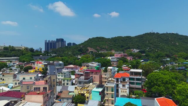 Aerial flyover reveals colorful rooftops of Coloane Village nestled among green hills with Cotai skyscrapers and casino towers on horizon