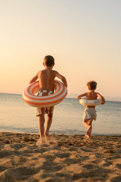 Two children holding inflatable swim rings on a white sandy beach at sunset, enjoying school holidays by the sea. A warm summer vacation scene filled with childhood joy, freedom and coastal memories.