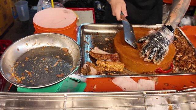 Street food vendor slicing crispy roasted pork belly on a wooden chopping block at a busy Thai night market. Traditional Thai street food preparation with crunchy pork crackling and fresh roasted meat