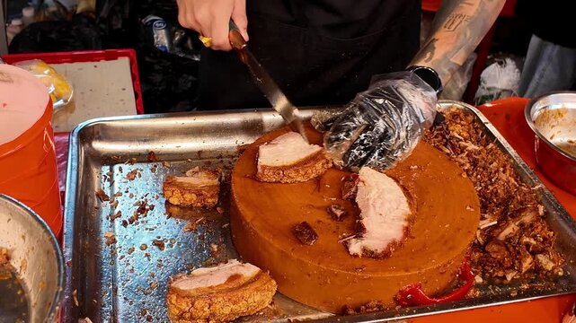 Close-up of a Thai street food vendor chopping crispy roasted pork belly with a cleaver on a large wooden chopping block at a busy night market. Traditional Thai street food preparation with crunchy p