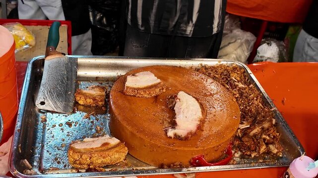 Close-up of a Thai street food vendor chopping crispy roasted pork belly into bite-sized pieces with a cleaver on a large wooden chopping block at a busy night market. Authentic Asian street food prep