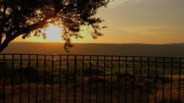 Sunset viewed from a lookout point over a Mediterranean landscape with trees and rolling hills, and a wrought-iron fence in the foreground, in Provence, southern France. 