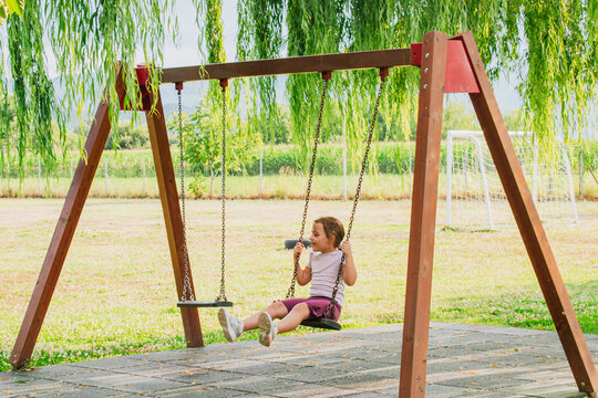 Side view of cute little girl swinging high on wooden swing set at outdoor park playground under green willow branches during warm sunny summer day
