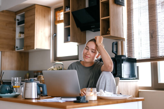 Stressed young woman working remotely on laptop at kitchen table, experiencing digital fatigue, emotional exhaustion and productivity pressure while working from home.