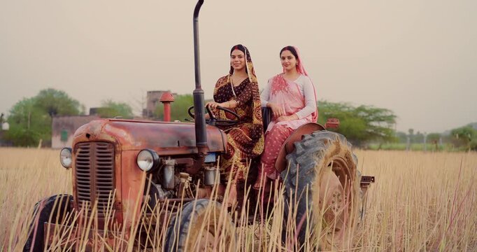 Indian young adult two woman sitting on tractor learn driving steering wheel look camera at farm rural area. Happy female wear saree staring cam do casual pose enjoy desi life day village ground place