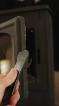 Vertical video. POV hand opens a wood-burning stove door revealing glowing embers and flames.