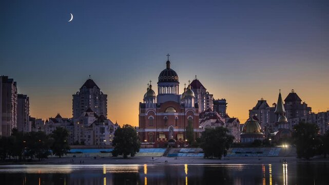 Young crescent moon setting after sunset above the Cathedral of the Nativity of Christ on Obolon Embankment in reflected in the calm waters of the Dnipro River