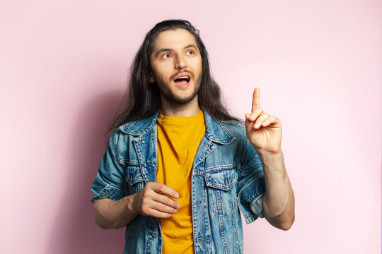Young happy man with long hair point with index finger up. Isolated on pastel pink background.