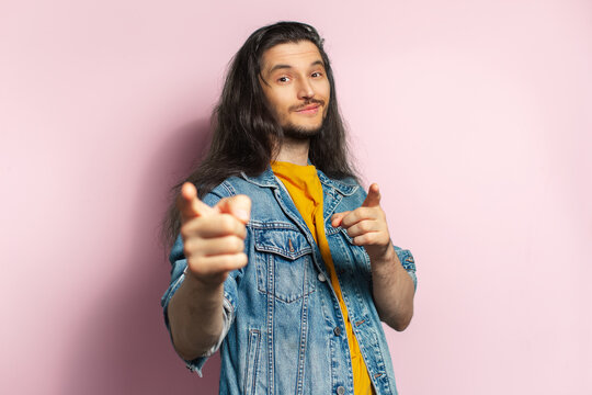 Young happy man with long hair point with index fingers on you. Isolated on pastel pink background.