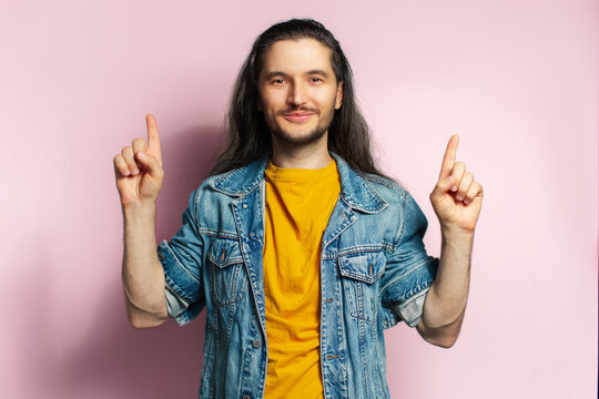 Young happy man with long hair point with fingers up. Isolated on pastel pink background.