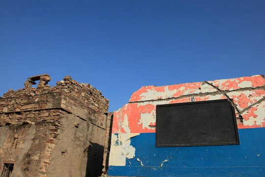 Weathered stone ruins beside a faded colorful wall with a blank black signboard under a clear blue sky