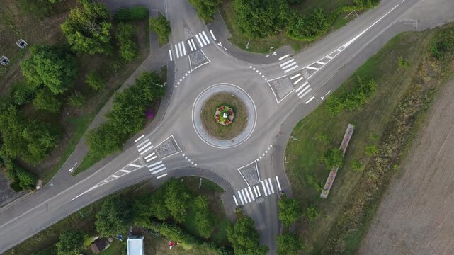 Aerial view of a roundabout in Dambach-La-Ville, France. A car drives through. The roundabout is surrounded by trees and grass, shot by a drone.
