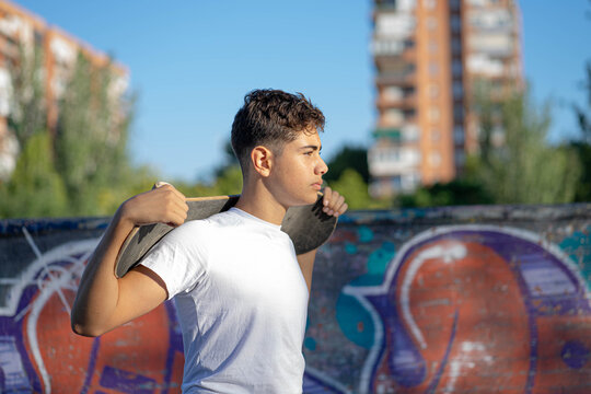 A serious boy walks with a skateboard resting on his shoulders