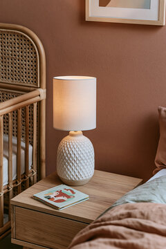 Boho nursery bedside area featuring a rattan crib, ceramic lamp, and illustrated book on a natural wood nightstand with warm wall colors