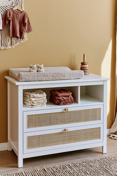 White changing dresser with rattan fronts and baby clothing in a boho nursery against a warm mustard yellow wall