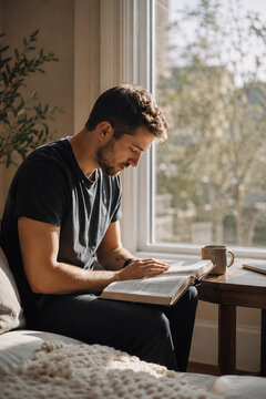 Young Christian man reading the Bible while sitting beside a sunlit window at home, personal faith, Bible study and morning devotion concept.