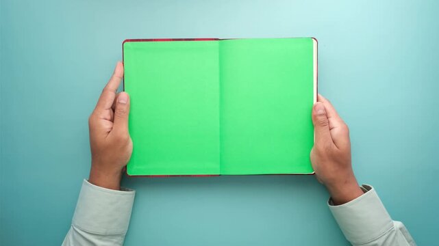 Hands holding open book with green screen pages over blue background for chroma key placement