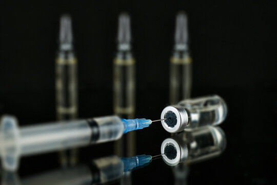 Syringe needle inserted into a glass vial with transparent liquid on a dark reflective surface. Concept of vaccination, medicine, healthcare, and medical treatment