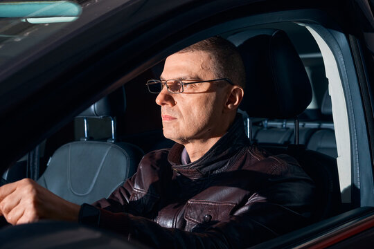 Serious mature man in glasses driving a car at night, side view through the window