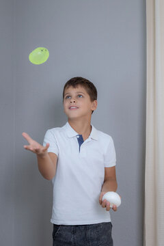 Young boy in white polo tossing bright water balloon indoors while holding another balloon, smiling with focused energy beside neutral gray wall in soft natural light for playful family lifestyle use