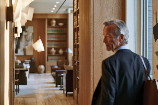 A reflective businessman with neat hair walks through a warm-toned corridor filled with elegant furnishings, showcasing confidence in a nuanced professional setting.