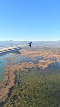 Airplane Window View Over Lake and Mountains. View from airplane window over lake, wetlands and mountains during flight. Aircraft wing, blue sky and aerial landscape from passenger seat.