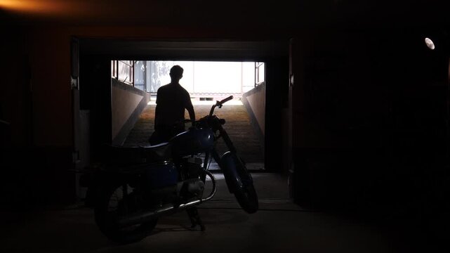 Man carrying a toolbox walking out of a dimly lit garage space towards a classic 1970s motorcycle, creating a strong silhouette against the bright outdoor light