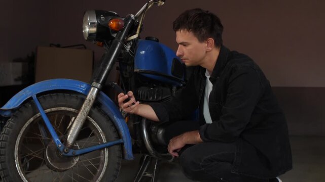 Man crouching analyzing a blue vintage motorcycle, using a smartphone for reference while working on a restoration project in a home garage