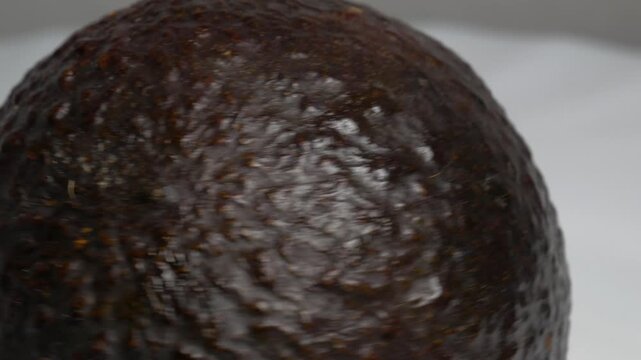 Close-up of a dark Hass avocado rotating on a white plate, showcasing its bumpy, textured skin and oval shape under bright studio lighting.
