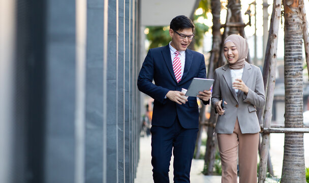 Young business muslim woman and business man colleague walking and discussing outside the office. Woman is holding a coffee and businessman is holding a digital tablet outdoor.