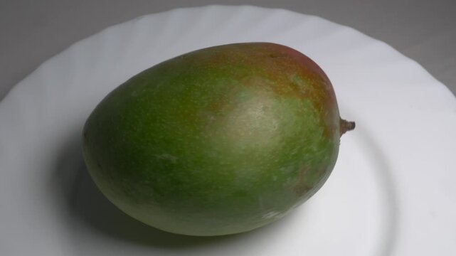 Close-up of a whole fresh green mango rotating on a white plate, showcasing its smooth skin, oval shape, and red blush under bright studio lighting.
