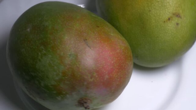 Close-up of a whole fresh green mango rotating on a white plate, showcasing its smooth skin, oval shape, and red blush under bright studio lighting.