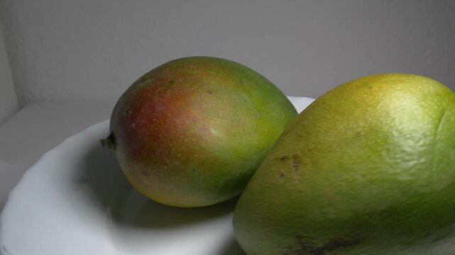 Close-up of a whole fresh green mango rotating on a white plate, showcasing its smooth skin, oval shape, and red blush under bright studio lighting.