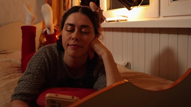 Beautiful young Caucasian woman smiling while using a smartphone lying on a bed at home in the evening. Social media, online communication, digital lifestyle, and cozy home relaxation concept.