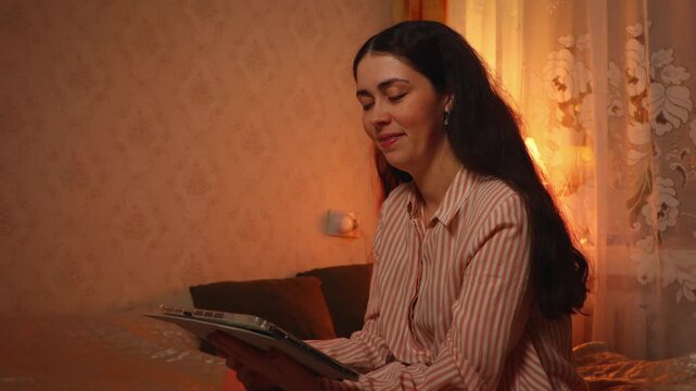 Side view of a young Caucasian woman relaxing and smiling while watching an online video on a tablet in the evening. Digital entertainment, home leisure, and modern lifestyle concept.