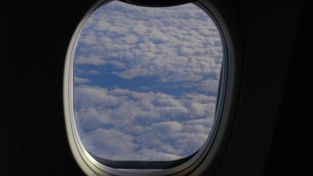 Looking through a round airplane window at the dense layer of white clouds floating over the blue sky at high altitude.
