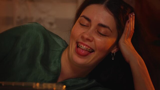 Portrait of a beautiful young Caucasian woman with braces laughing and smiling while using a smartphone, tablet, or reading a book at home in the evening. Digital lifestyle, entertainment