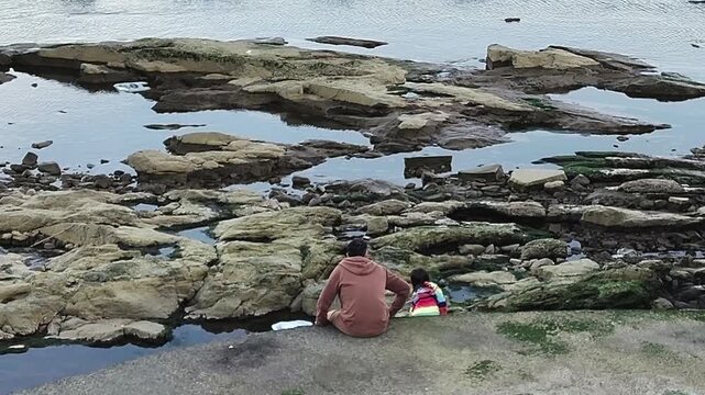 Long shot video with high angle and fixed camera showing a father and his five-year-old daughter sitting on the edge of a rocky beach, montevideo city, uruguay
