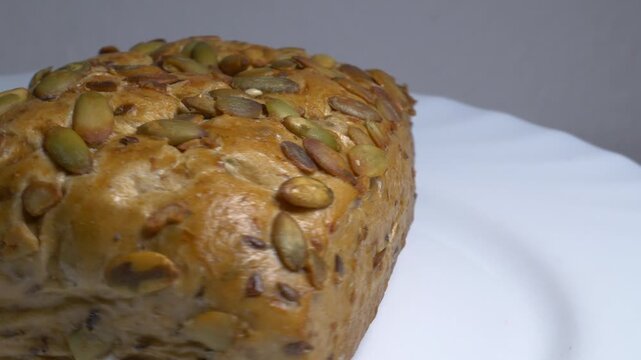 Close-up of a fresh whole grain bread roll with pumpkin seeds rotating on a white plate, showcasing its golden crust and textured surface under studio lights.