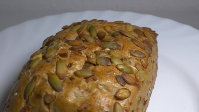 Close-up of a fresh whole grain bread roll with pumpkin seeds rotating on a white plate, showcasing its golden crust and textured surface under studio lights.