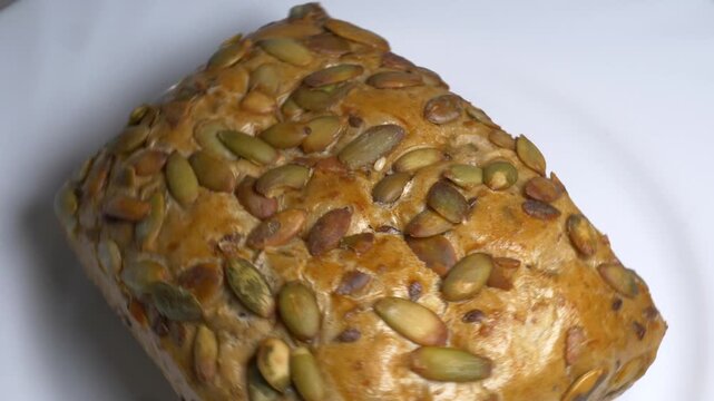 Close-up of a fresh whole grain bread roll with pumpkin seeds rotating on a white plate, showcasing its golden crust and textured surface under studio lights.