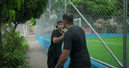 Two Hispanic men outdoors by a soccer field greeting each other warmly, talking and embracing with...